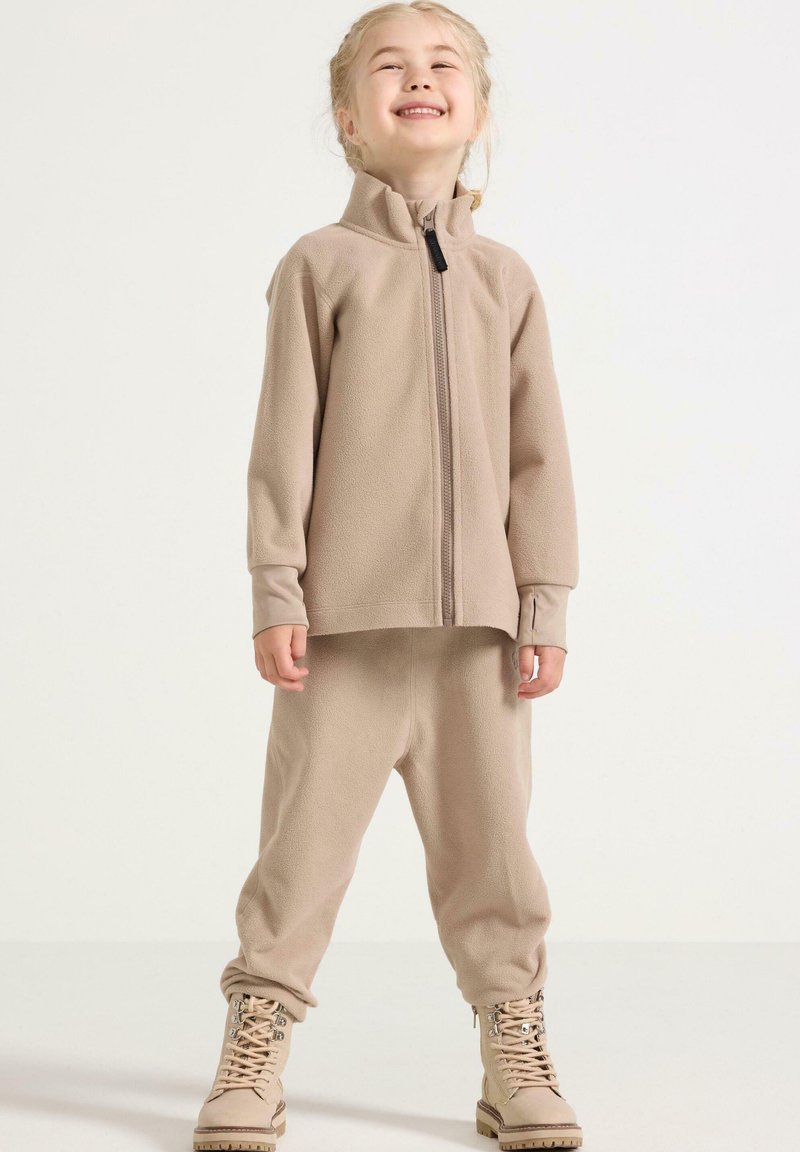 Completo in pile beige con giacca con zip e pantaloni abbinati. La giacca presenta un colletto alto, mentre i pantaloni hanno una vestibilità comoda. Stivali beige completano il look.