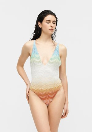 Femeie cu păr ud purtând un costum de baie întreg în degrade multicolor, cu decolteu adânc în V, stând în fața unui fundal gri deschis.