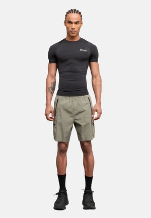 Atleta con capelli intrecciati che indossa una maglietta nera aderente con la scritta "Utopia", pantaloncini cargo beige, calzini neri e scarpe da ginnastica, in piedi su sfondo bianco.