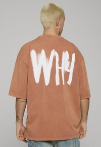 Oversized oranje-bruin T-shirt met "WHY" in het wit gespoten op de achterkant. Gemaakt van katoen, korte mouwen, losse pasvorm en een ronde hals.