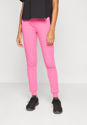 Person, die knallpinke, eng anliegende Jogginghosen und schwarze Turnschuhe trägt, vor einem schlichten hellgrauen Hintergrund steht.