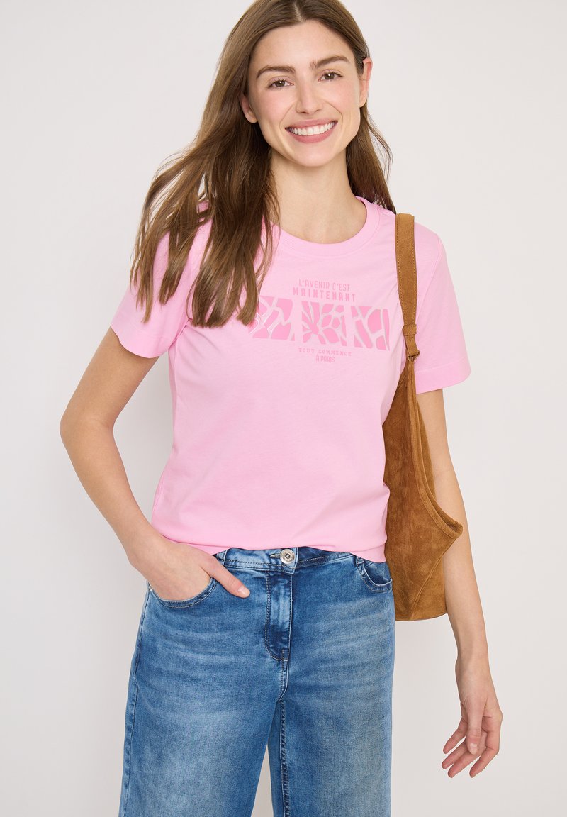 Ung kvinde smilende, iført en lyserød t-shirt med grafik, blå jeans og en brun skuldertaske mod en ensartet lys baggrund.