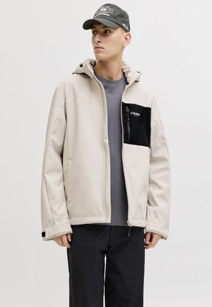 Mies, jolla on beige takki mustalla rintataskulla, harmaa paita, mustat housut ja musta lippalakki, katsoo sivulle yksiväristä taustaa vasten.