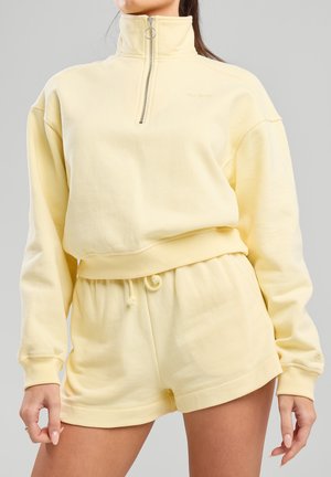 Gelber Baumwollpullover mit hohem Kragen und Viertel-Reißverschluss, kombiniert mit passenden Shorts mit elastischem Bund. Glatte Textur.