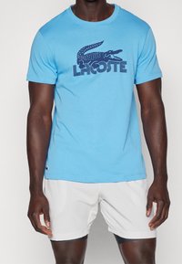Man wearing a blue Lacoste T-shirt with a large crocodile logo and white athletic shorts against a plain background.