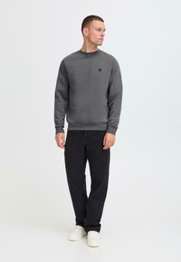 Felpa grigia con collo rotondo e polsini a coste, abbinata a jeans neri e sneakers bianche. Presenta un piccolo logo nero sul petto.