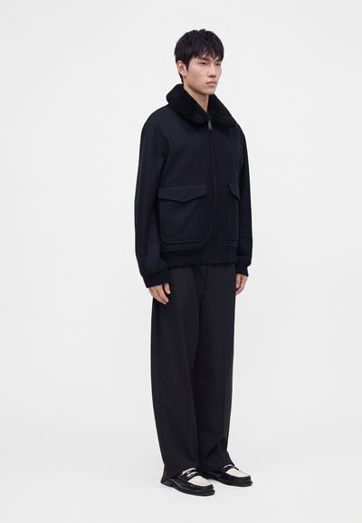 Jeune homme portant une veste noire zippée avec un col en fourrure, un pantalon noir ample et des mocassins noirs et blancs, debout devant un fond uni.