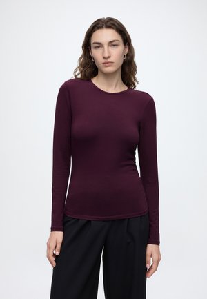 Femme aux cheveux bruns ondulés portant un haut ajusté à manches longues violet foncé et un pantalon noir ample sur un fond clair uni.
