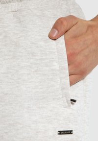 Hellgraue Jogginghosen aus einem strukturierten Stoff, mit einer Seitentasche und einem schwarzen Akzent-Logo-Tag nahe der Taschenöffnung.