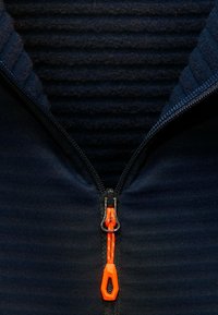 Dunkelblaue Jacke mit Reißverschluss und gerippter Textur unter dem Reißverschluss. Oranger Reißverschlussgriff mit schwarzem Zubehör. Weicher Stoff, Nahaufnahme.