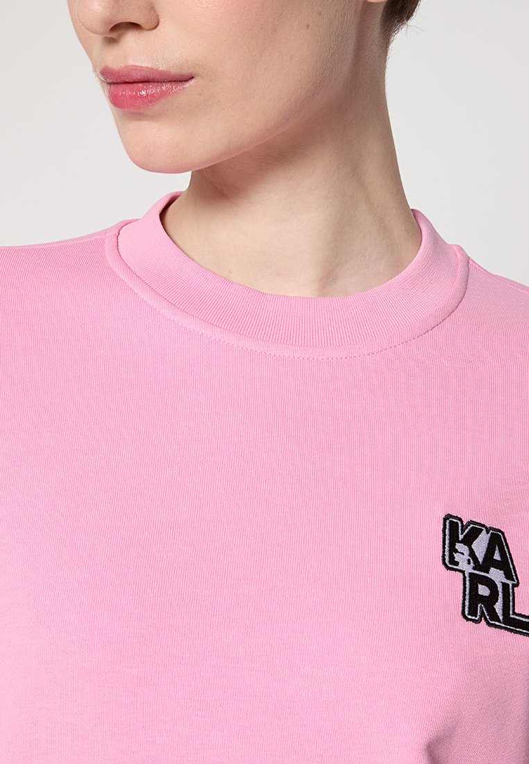 Nahaufnahme einer Person, die ein pinkes, geripptes Rundhalsausschnitt-Shirt mit einem kleinen schwarz-weißen "KARL"-Logo auf der Brust trägt.