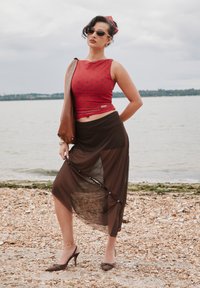 Crveni top bez rukava s točkicama, smeđa prozirna suknja i smeđe cipele s visokim potpeticama. Dodaci uključuju tamne sunčane naočale i smeđu torbu.