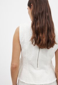White textured sleeveless top with a distinct pattern, featuring a center back zipper and a curved hemline.