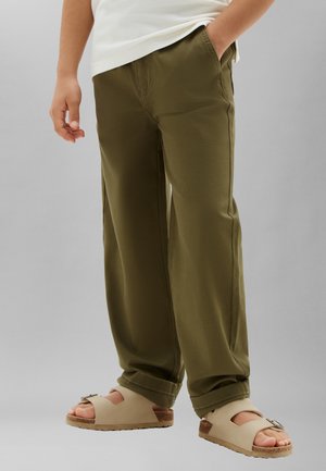 Personne portant un pantalon vert olive, un haut blanc et des sandales beige à boucle, debout avec une main dans la poche sur un fond neutre.