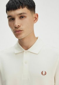 Polo bianco con una trama testurizzata, dotato di un logo ricamato marrone sul petto sinistro e di due bottoni al colletto.