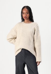 Cremfarbener, grob gestrickter Pullover mit lockerer Passform, weiten Bündchen und einem High-Low-Saumdesign, kombiniert mit dunklen, weit geschnittenen Hosen.