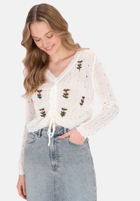 Fehér, kötött crop top virágmintás hímzéssel, V kivágással, megkötős elülső résszel és nyitott horgolási mintákkal. Világoskék farmerral kombinálva.