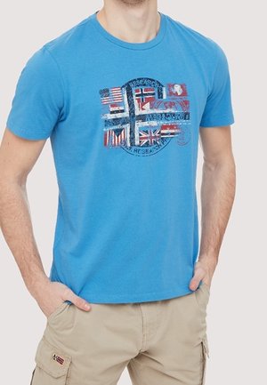Mann trägt ein blaues T-Shirt mit mehreren internationalen Flaggen und beige Cargoshorts mit einem kleinen Flaggenaufnäher an der Tasche.