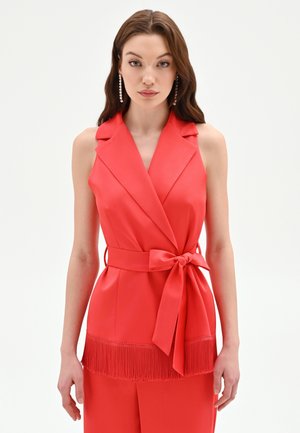 Femme portant un haut cache-cœur sans manches rouge avec col, nœud à la taille et détails à franges, assorti d'un pantalon et de boucles d'oreilles pendantes.