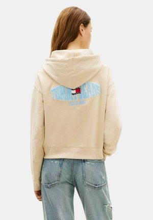 Donna con felpa beige con cappuccio con logo "Tommy Jeans" azzurro chiaro e jeans chiari strappati, di spalle contro uno sfondo neutro.