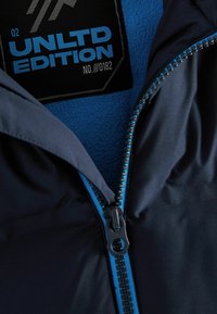 Marinejakke med en blå lynlås, som har et stofmærke med teksten "UNLTD EDITION NO. 0182" på en struktureret blå foring.