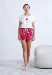 Λευκό βαμβακερό T-shirt με την επιγραφή "AMOUR" σε κόκκινο, συνδυασμένο με κόκκινα σορτς που έχουν άσπρα σχέδια με καρδιές. Το μοντέλο φοράει μπεζ παντόφλες και στέκεται σε μαλακή επιφάνεια.