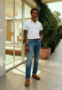 Hvit poloshirt, blå denimjeans og brune sko. Står utendørs foran en moderne designbakgrunn med planter og naturlig lys.