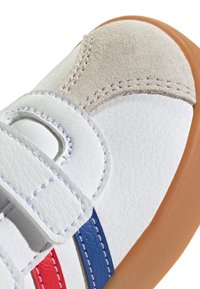 Zapatilla de cuero blanco con rayas azules y rojas, con un acento de ante gris texturizado y suela de goma color tostado. Cierre con tira de velcro.