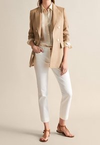 Beżowa dwurzędowa marynarka, kremowa koszula i białe spodnie o obciętej długości. Brązowe płaskie sandały dopełniają strój. Prosty i dopasowany krój.