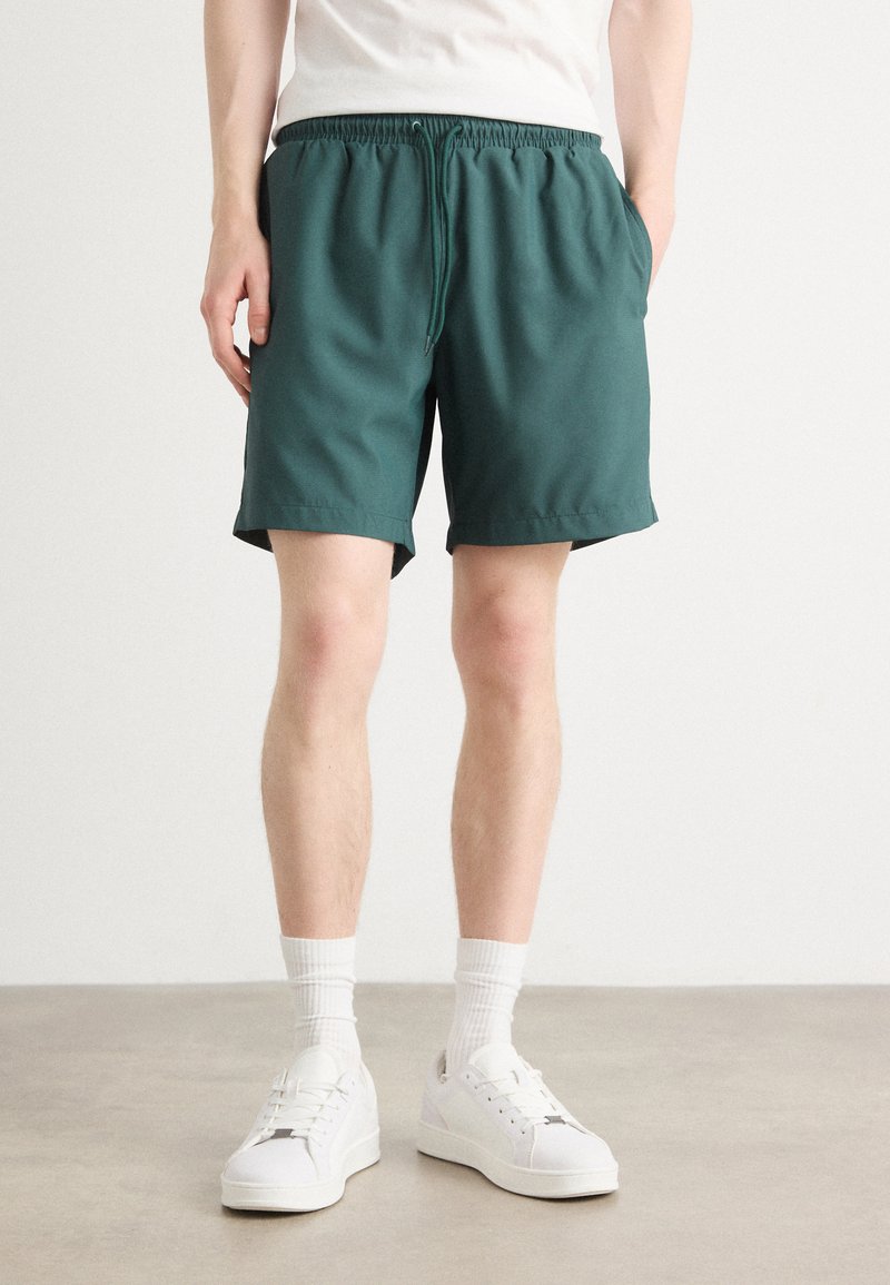 Shorts vert avec taille élastique et poches latérales, en tissu lisse, longueur au-dessus du genou, associés à des chaussures et des chaussettes blanches.