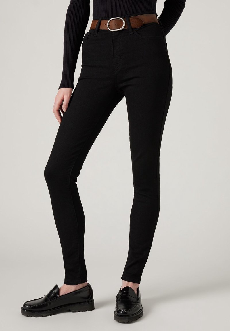 Jean skinny noir taille haute en mélange de coton extensible, avec une ceinture en cuir marron et une boucle en métal argenté au design épuré.
