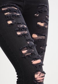 Jeans negros rasgados con bordes deshilachados, que presentan agujeros irregulares que revelan una tela más clara debajo. Vista en primer plano de las áreas remendadas.