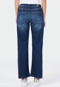 Persoon in donkerblauwe rechte spijkerbroek met achterzakken en een wit topje, staande voor een effen lichte achtergrond.