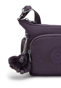 Lila Stoff-Crossbody-Tasche mit einem Reißverschlussfach, das ein rundes Logo und einen pelzigen Gorilla-Schlüsselanhänger zeigt. Verstellbarer Tragegurt mit Hardware-Accents.