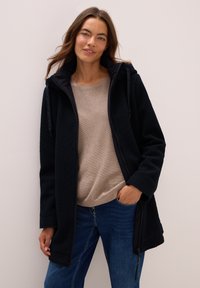 Zwarte fleece jas met een ritssluiting aan de voorkant, capuchon en twee zijzakken. Draag het over een lichtbruine gestructureerde trui en blauwe jeans.