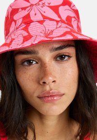 Červený klobúk s kvetinovým vzorom v ružovej farbe, vyrobený z tkaniny s textúrovaným povrchom. Na prednej strane sa nachádza vyšité logo značky.