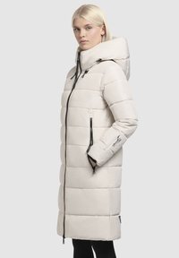 Manteau rembourré beige clair avec capuche, doté d'une fermeture à zip sur le devant, de poches latérales zippées et d'une texture matelassée. Cordons réglables présents.