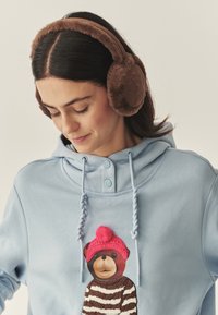 Barna műszőrme fülvédő kör alakú kialakítással, világoskék kapucnis pulóverrel párosítva, amelyen egy rózsaszín sapkás, csíkos pulóveres maci rajzfilmfigura látható.