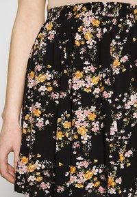 Jupe noire à fleurs avec des plis, présentant des fleurs roses et orange, taille élastique, et texture de tissu douce. Vue en gros plan avec une main.