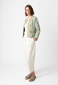 Uzorčana zelena jakna s narančastim naglascima, okrugli izrez, kopčanje na gumbi i dugi rukavi. Stilizirana preko kremaste bluze i širokih hlača.