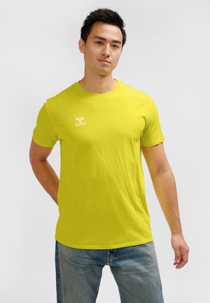 Mężczyzna ubrany w jaskrawożółty t-shirt z krótkim rękawem i małym białym logo hummel, w połączeniu z niebieskimi jeansami, stojący na białym tle.