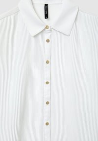 Camicia bianca con bottoni a punta realizzata in tessuto plissettato, con dettagli dei bottoni dorati lungo la parte anteriore.