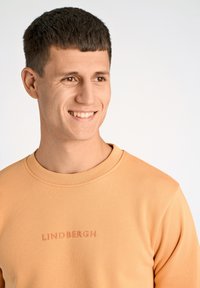 Orange stickad sweatshirt med ribbat rundhals. Har en subtil präglad "LINDBERGH"-logga på bröstet. Slät textur, avslappnad passform.