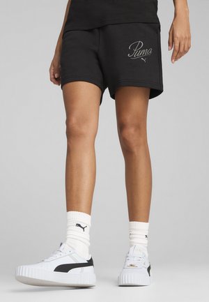 Pantalones cortos de algodón negros con logo bordado de Puma, de longitud por encima de la rodilla, combinados con zapatillas blancas con detalles en negro y calcetines blancos de canalé.