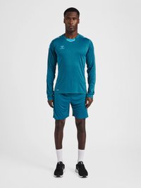 Teal langærmet atletisk top med kontrastfarvet krave, parret med teal shorts. Sorte sneakers og hvide sokker fuldender outfittet.