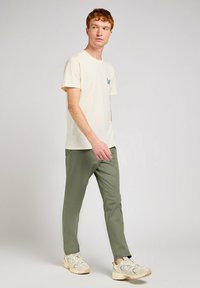 Ljus krämfärgad bomulls t-shirt med tealfärgad logoficka, parat med slim-fit olivgröna byxor och beige sneakers med grå detaljer.
