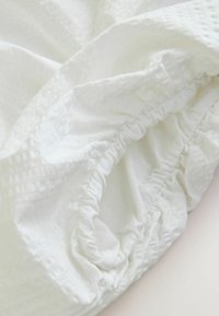 Coin de drap-housse blanc avec bande élastique fronçée et tissu texturé sur une surface claire.