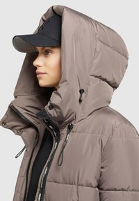 Padded winter jacket in taupe, featuring a large hood, zip closure, and drawstrings. Black cap with logo complements the look.