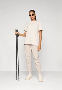 Beigā T-kreklā ar sarkano logo, saskaņotās beigu bikses un gaišu krāsu sporta apaviem. Tur rokās melnas pārgājiena nūjas, valkā gaismas saulesbrilles.