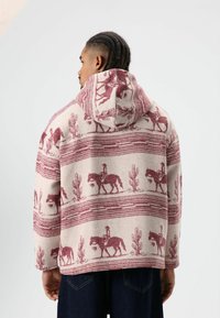 Homme aux cheveux tressés portant un sweat à capuche beige orné de motifs de cow-boys bordeaux, de chevaux et de cactus, debout devant un fond blanc uni.